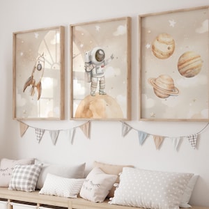 Puede incluir: Tres impresiones de acuarela enmarcadas con un tema espacial. Las impresiones presentan un astronauta, un cohete y planetas. Las impresiones están colgadas en una pared blanca sobre un banco de madera con cojines.