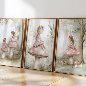 May include: Set of three framed art prints featuring watercolor illustrations of fairies. Each print depicts a fairy in a pink dress sitting on a mushroom in a forest setting. The frames are a warm, light brown color.