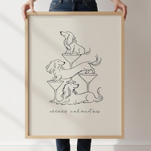Può includere: Una stampa artistica incorniciata con un disegno a linee nere di tre cani bassotti che interagiscono con bicchieri da martini su uno sfondo beige chiaro. Il testo "weenies and martinis" è stampato in un semplice carattere corsivo nero sotto l'illustrazione.