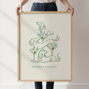 May include: A framed vertical art print on a cream background featuring a line art illustration of three dachshunds interacting with martini glasses. The text reads WEENIES & MARTINIS and GOOD DRINKS, BETTER COMPANY in dark green lettering.