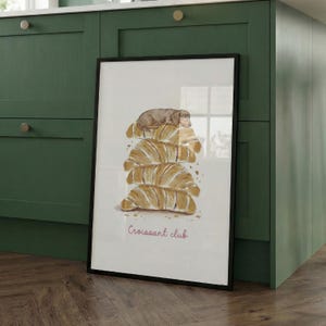 Puede incluir: Una l&aacute;mina enmarcada con una ilustraci&oacute;n en acuarela de un perro salchicha marr&oacute;n descansando sobre una pila de cuatro cruasanes dorados. El texto "Croissant club" est&aacute; escrito en una fuente cursiva rosa debajo de la ilustraci&oacute;n sobre un fondo blanco.