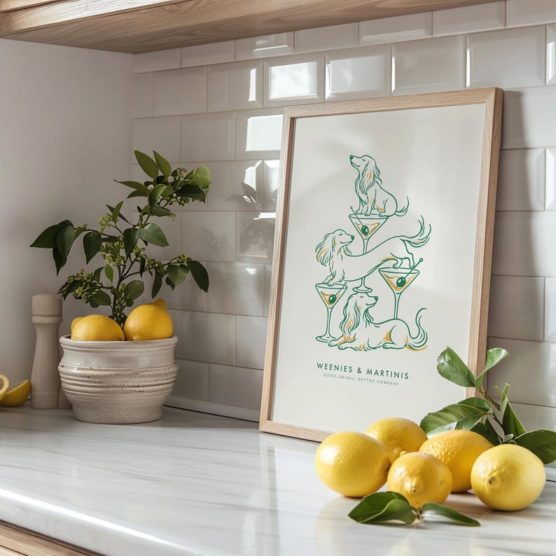 May include: A framed vertical art print featuring a green line drawing of three dachshund dogs balanced on martini glasses. The text reads WEENIES & MARTINIS GOOD DRINKS, BETTER COMPANY. The print is displayed in a light wood frame on a white surface.