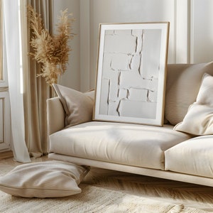 May include: A framed abstract art print with a textured, off-white design, resting on a beige sofa. The artwork features a series of rectangular shapes and lines. The room has a neutral color palette with a beige rug and pillows.