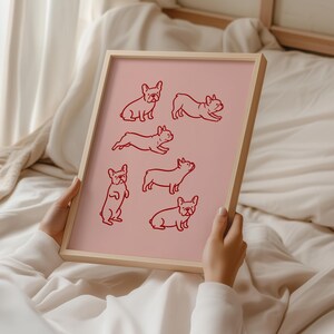 May include: A framed pink print featuring red line drawings of French Bulldogs in various poses. The artwork is held by a person, showcasing the print's light wooden frame. The background is a soft, neutral-toned fabric.