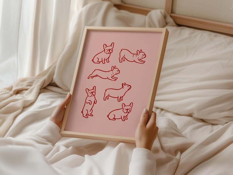May include: A framed pink print featuring red line drawings of French Bulldogs in various poses. The artwork is held by a person, showcasing the print's light wooden frame. The background is a soft, neutral-toned fabric.
