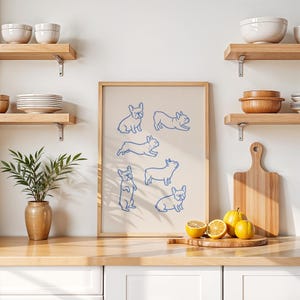 May include: A framed print featuring blue line drawings of French Bulldogs in various poses. The artwork is displayed on a kitchen counter with lemons, a cutting board, and wooden shelves with bowls and plates.