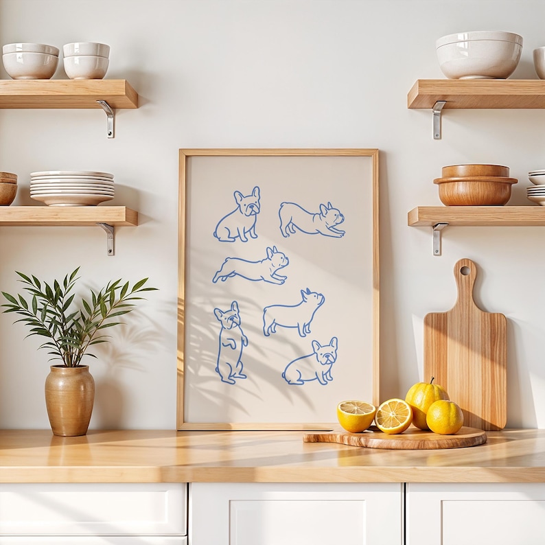May include: A framed print featuring blue line drawings of French Bulldogs in various poses. The artwork is displayed on a kitchen counter with lemons, a cutting board, and wooden shelves with bowls and plates.