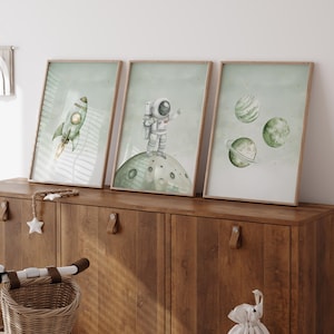 Pu&ograve; includere: Tre stampe incorniciate ad acquerello con illustrazioni a tema spaziale. Una mostra un razzo verde con una fiamma gialla, un'altra un astronauta su una luna e la terza, pianeti. Le stampe hanno uno sfondo verde chiaro e sono in cornici di legno.