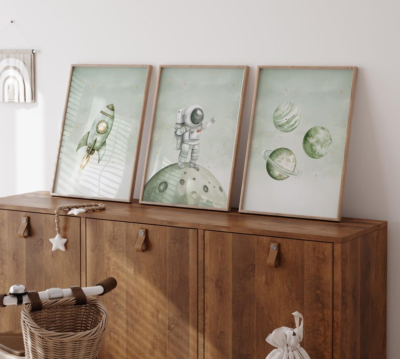 Pu&ograve; includere: Tre stampe incorniciate ad acquerello con illustrazioni a tema spaziale. Una mostra un razzo verde con una fiamma gialla, un'altra un astronauta su una luna e la terza, pianeti. Le stampe hanno uno sfondo verde chiaro e sono in cornici di legno.