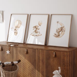 May include: Three framed art prints featuring watercolor illustrations of bunnies. One shows a bunny in a hot air balloon, another with a balloon on a cloud, and the third on a crescent moon. The frames are light wood, and the art has a soft, neutral color palette.