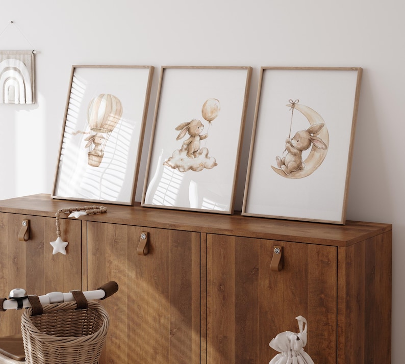 May include: Three framed art prints featuring watercolor illustrations of bunnies. One shows a bunny in a hot air balloon, another with a balloon on a cloud, and the third on a crescent moon. The frames are light wood, and the art has a soft, neutral color palette.