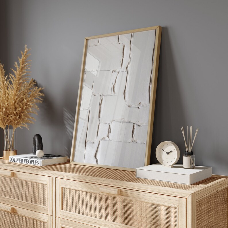 May include: A framed abstract art piece with a textured white surface leans against a gray wall. The artwork is displayed on a light wood cabinet with a woven cane design, alongside decorative items like a clock and a reed diffuser.