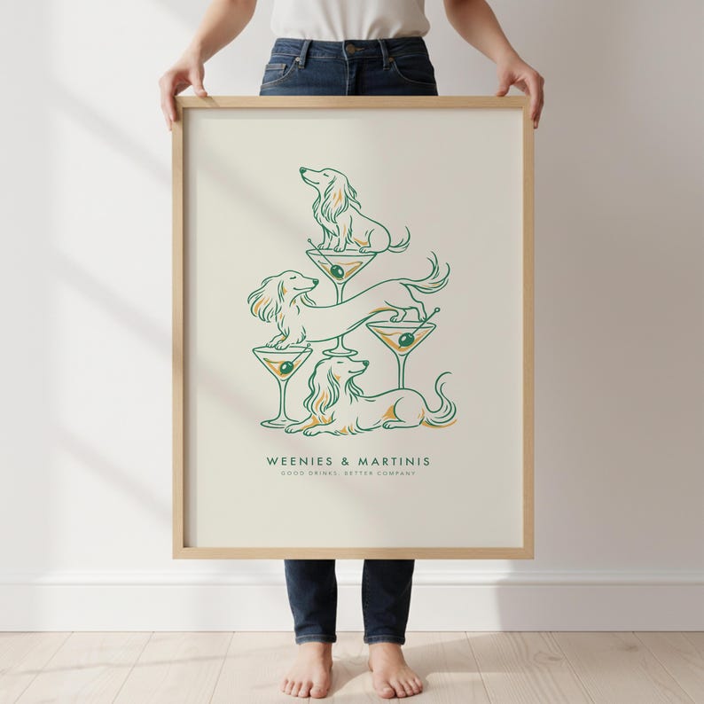 May include: A framed vertical art print on a cream background featuring a line art illustration of three dachshunds interacting with martini glasses. The text reads WEENIES & MARTINIS and GOOD DRINKS, BETTER COMPANY in dark green lettering.