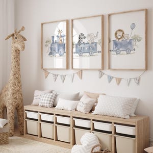 May include: Three framed wall art prints featuring watercolor illustrations of animals riding in blue train cars. The first shows a giraffe, the second a zebra and elephant, and the third a lion and bear with a blue balloon. A bunting banner hangs below.