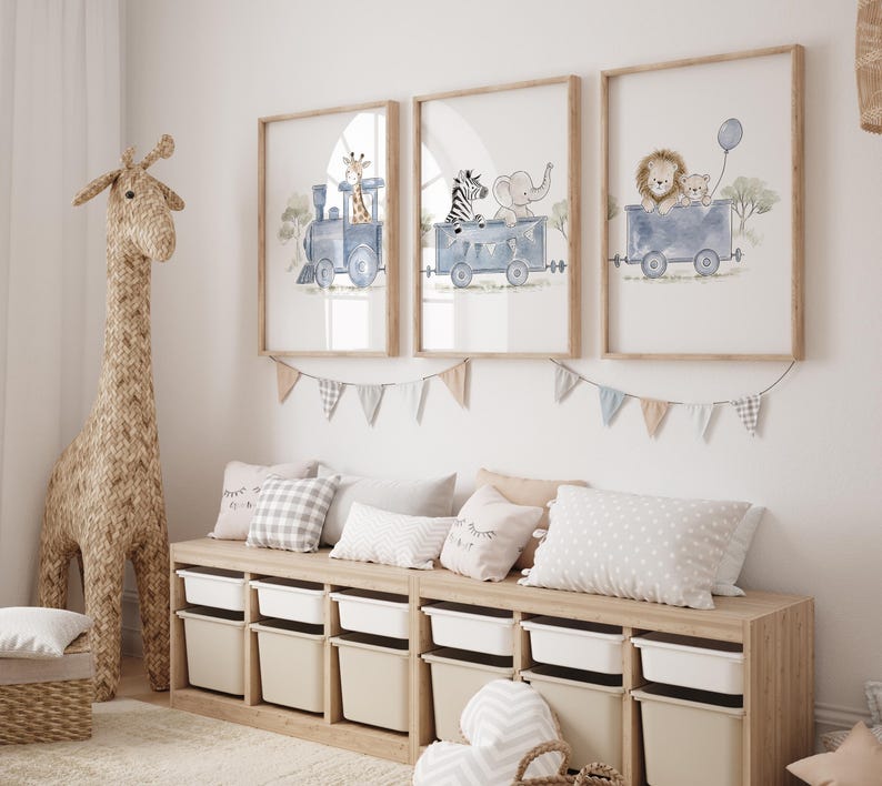 May include: Three framed wall art prints featuring watercolor illustrations of animals riding in blue train cars. The first shows a giraffe, the second a zebra and elephant, and the third a lion and bear with a blue balloon. A bunting banner hangs below.