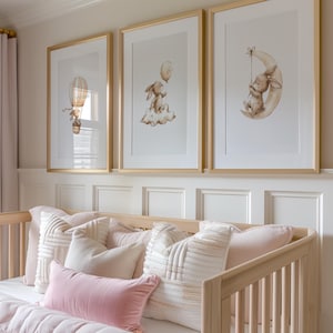 May include: Three framed art prints with gold frames, featuring watercolor illustrations of a bunny with a balloon, a bunny on a cloud, and a bunny on a crescent moon. The prints hang above a light wood crib with pink and white pillows and a pink blanket.