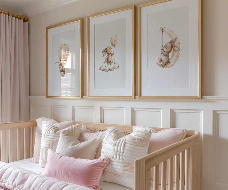 May include: Three framed art prints with gold frames, featuring watercolor illustrations of a bunny with a balloon, a bunny on a cloud, and a bunny on a crescent moon. The prints hang above a light wood crib with pink and white pillows and a pink blanket.
