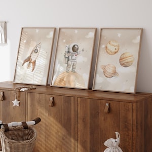 Pu&ograve; includere: Tre stampe artistiche incorniciate ad acquerello con illustrazioni a tema spaziale. Le stampe raffigurano un razzo, un astronauta e pianeti su uno sfondo beige con nuvole e stelle. Le cornici sono di colore marrone chiaro e l'opera &egrave; esposta su un mobile di legno.