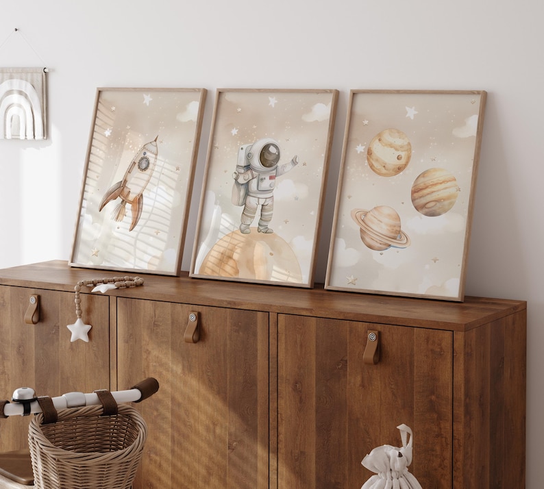 Pu&ograve; includere: Tre stampe artistiche incorniciate ad acquerello con illustrazioni a tema spaziale. Le stampe raffigurano un razzo, un astronauta e pianeti su uno sfondo beige con nuvole e stelle. Le cornici sono di colore marrone chiaro e l'opera &egrave; esposta su un mobile di legno.