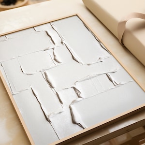 May include: A framed abstract art piece with a textured white surface. The artwork features a series of rectangular and angled shapes, creating a layered effect. The frame is made of light-colored wood, adding a natural touch to the modern design.