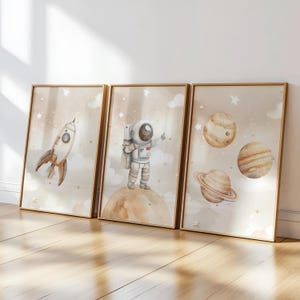 Pu&ograve; includere: Tre stampe artistiche incorniciate con illustrazioni a tema spaziale. Una mostra un razzo, un'altra un astronauta e la terza raffigura pianeti. L'opera d'arte ha uno stile morbido ad acquerello con una tavolozza di colori neutri.