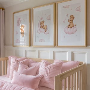 K&ouml;nnte beinhalten: Drei gerahmte Kunstdrucke mit goldenen Rahmen, die jeweils eine skurrile Illustration eines Baby-Tiers auf einer Wolke zeigen. Die Drucke befinden sich in einem Kinderzimmer, &uuml;ber einem hellen Holzbett mit rosa Bettw&auml;sche und Kissen.