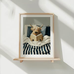 May include: A framed print of a Highland cow reading a book in bed. The cow is light brown with long hair and horns. The bed has a striped blanket and blue cushions. The artwork is displayed on a wooden shelf.