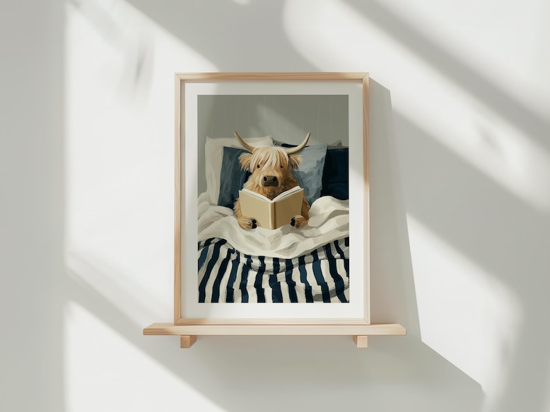 May include: A framed print of a Highland cow reading a book in bed. The cow is light brown with long hair and horns. The bed has a striped blanket and blue cushions. The artwork is displayed on a wooden shelf.