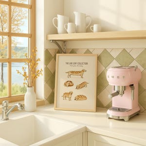 Puede incluir: Una l&aacute;mina enmarcada titulada THE LAB LOAF COLLECTION con ilustraciones de perros golden retriever con forma de hogazas de pan. La l&aacute;mina se muestra sobre una encimera de cocina junto a una cafetera espresso rosa pastel y un jarr&oacute;n blanco con flores amarillas.