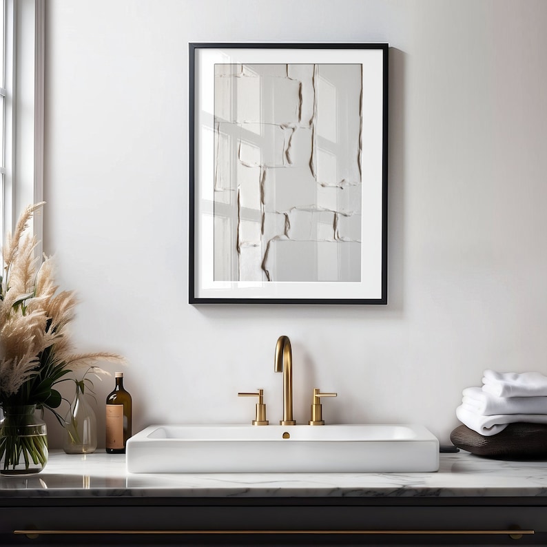 May include: A framed abstract art print with a textured, neutral-toned design hangs above a white rectangular sink. The artwork is in a black frame. A gold faucet and a vase of pampas grass add to the bathroom's decor.
