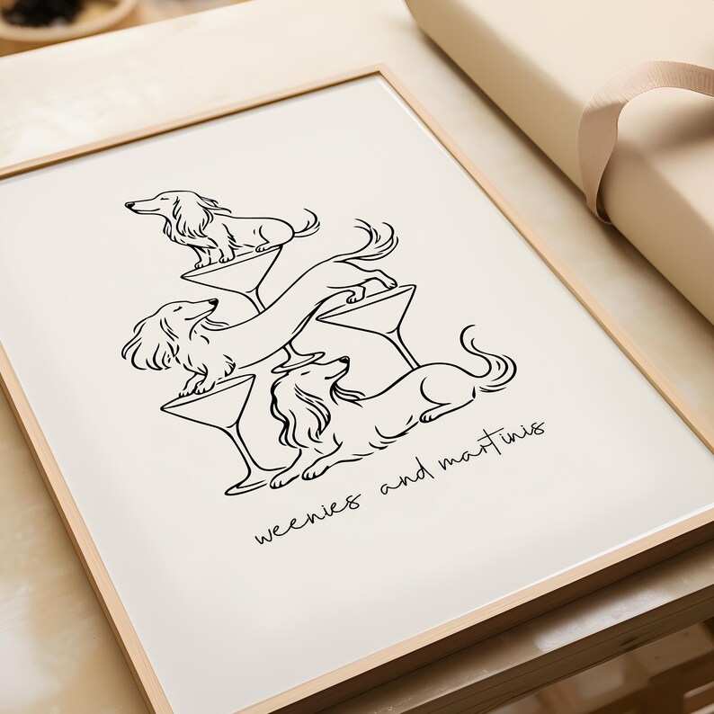 May include: A framed art print featuring a black line drawing of three dachshunds interacting with martini glasses. The dogs are positioned in a stacked arrangement with the text weenies and martinis written in a cursive script below the illustration.