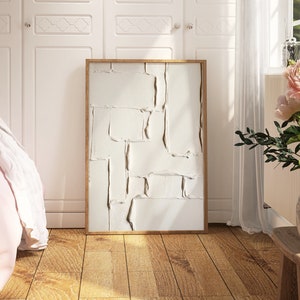 May include: A framed abstract art piece with a textured, off-white surface and a natural wood frame. The artwork is displayed in a bright room with a wooden floor, a bed, and a vase of pink flowers.