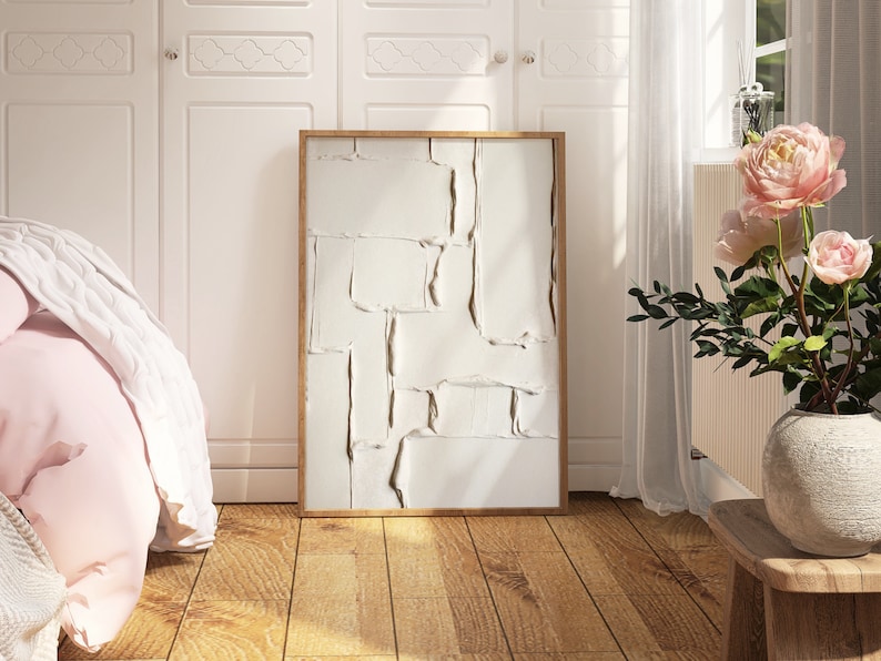 May include: A framed abstract art piece with a textured, off-white surface and a natural wood frame. The artwork is displayed in a bright room with a wooden floor, a bed, and a vase of pink flowers.