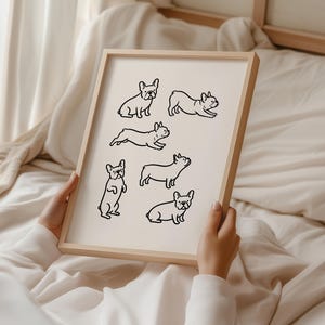 May include: A framed print featuring black line drawings of French Bulldogs in various poses. The dogs are depicted sitting, standing, and running on a cream-colored background. The frame is made of light-colored wood.