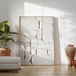 May include: An interior design scene featuring a large abstract artwork with a gold frame. A large potted plant, a gray ottoman, and terracotta vases are also present. The room has white paneling and a wooden floor.