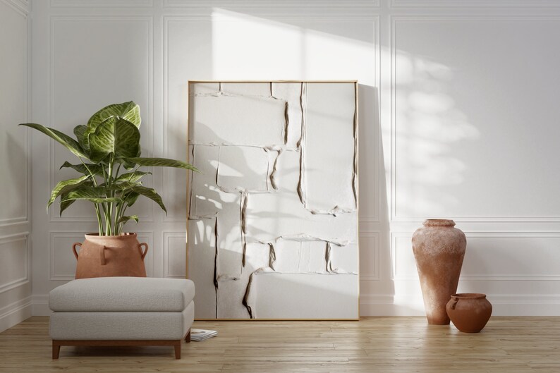 May include: An interior design scene featuring a large abstract artwork with a gold frame. A large potted plant, a gray ottoman, and terracotta vases are also present. The room has white paneling and a wooden floor.