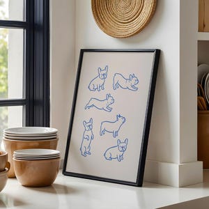 May include: A framed art print featuring blue line drawings of French Bulldogs in various poses. The artwork is displayed in a black frame and rests on a white countertop. The background includes a window, bowls, and a woven wall hanging.
