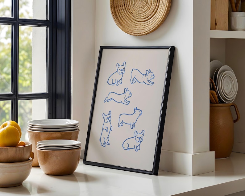 May include: A framed art print featuring blue line drawings of French Bulldogs in various poses. The artwork is displayed in a black frame and rests on a white countertop. The background includes a window, bowls, and a woven wall hanging.