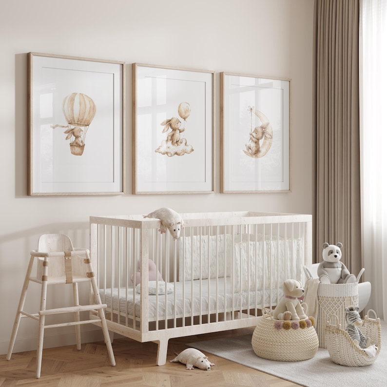 May include: A nursery scene featuring three framed watercolor prints with bunny illustrations. A white crib, high chair, and decorative baskets filled with stuffed animals complete the room's decor. The prints depict a bunny in a hot air balloon, on a cloud, and on a crescent moon.