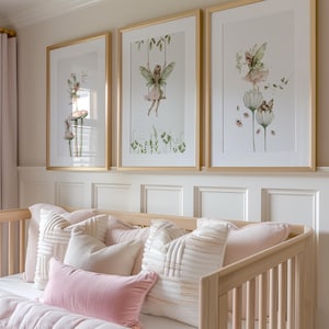 May include: Three framed art prints featuring watercolor illustrations of fairies in a nursery setting. The prints have gold frames and depict fairies on swings, flowers, and mushrooms. The nursery includes a light wood crib with pink and white pillows.