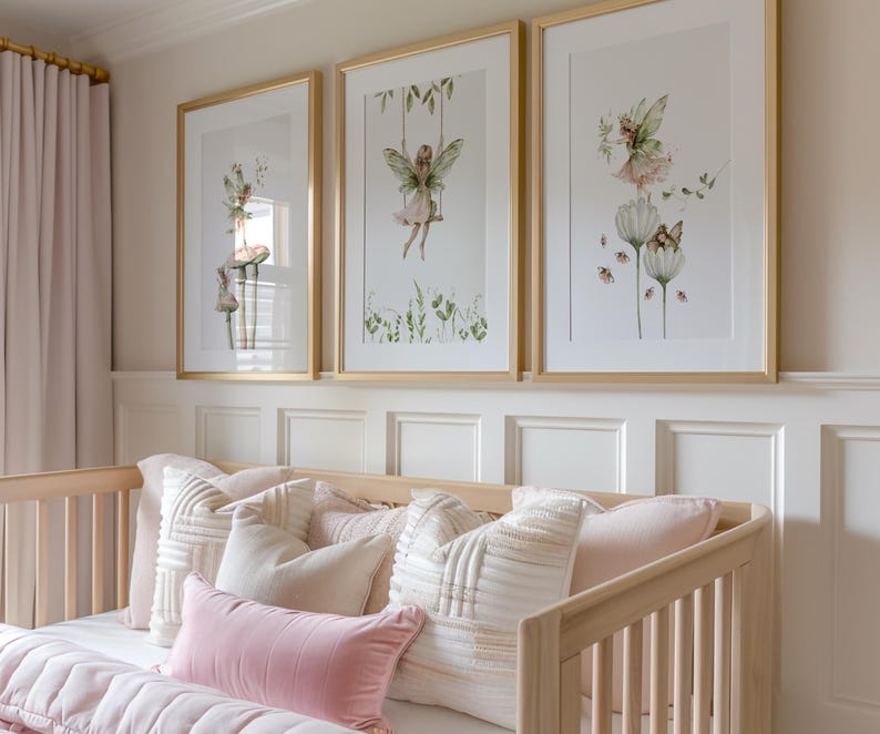 May include: Three framed art prints featuring watercolor illustrations of fairies in a nursery setting. The prints have gold frames and depict fairies on swings, flowers, and mushrooms. The nursery includes a light wood crib with pink and white pillows.