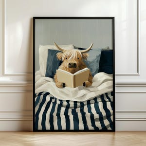 May include: A framed art print showing a Highland cow reading a book in bed. The cow is light brown with long, shaggy fur and horns. The bed has white and navy striped bedding and pillows. The artwork is in a black frame.