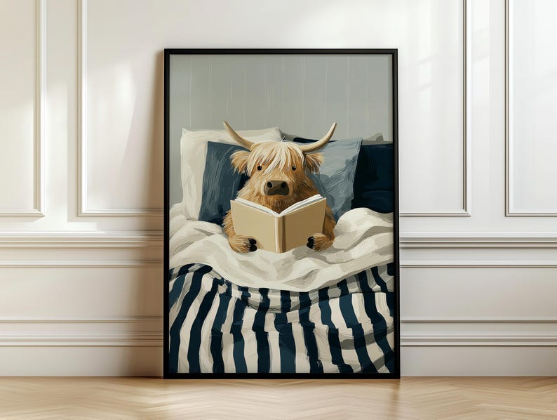 May include: A framed art print showing a Highland cow reading a book in bed. The cow is light brown with long, shaggy fur and horns. The bed has white and navy striped bedding and pillows. The artwork is in a black frame.