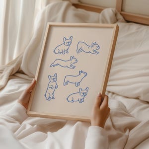 May include: A framed print featuring blue line drawings of French Bulldogs in various poses. The artwork is set against a cream-colored background and encased in a light wooden frame. The print is held against a backdrop of soft, neutral-toned fabric.