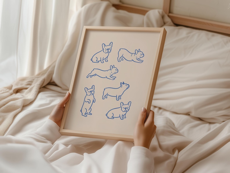 May include: A framed print featuring blue line drawings of French Bulldogs in various poses. The artwork is set against a cream-colored background and encased in a light wooden frame. The print is held against a backdrop of soft, neutral-toned fabric.