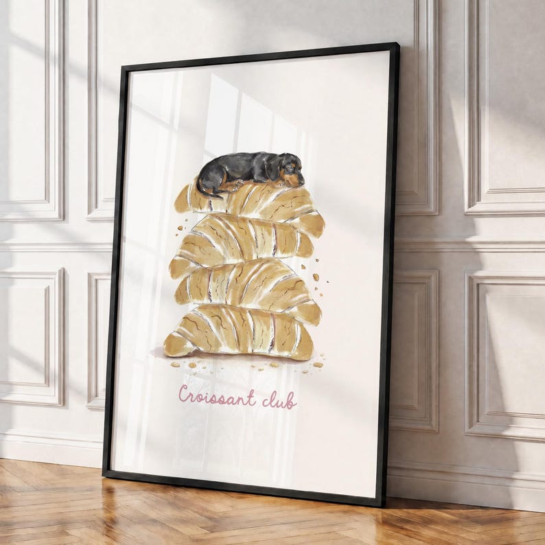 Puede incluir: Una l&aacute;mina de arte enmarcada con una ilustraci&oacute;n en acuarela de un perro salchicha negro y fuego descansando sobre una pila de cuatro cruasanes dorados. El texto "Croissant club" aparece en cursiva rosa bajo la pila de pasteles.