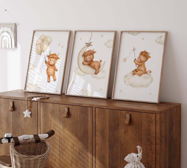 K&ouml;nnte beinhalten: Set aus drei gerahmten Aquarelldrucken mit einem Highland-Kuh-Thema. Ein Druck zeigt eine Kuh mit Luftballons, ein anderer auf einem Mond, und der dritte auf einer Wolke mit einem Stern. Die Drucke haben eine weiche, neutrale Farbpalette.