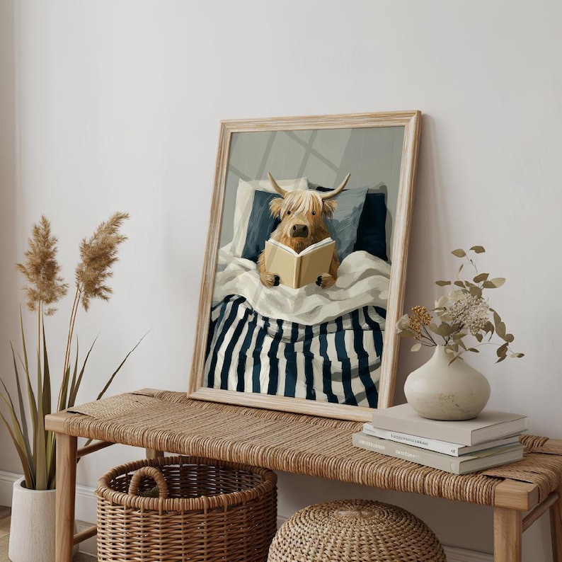 May include: A framed art print showing a Highland cow reading a book in bed. The cow has a shaggy, light brown coat and horns, under a white duvet and navy blue and white striped bedding. The artwork is presented in a light wood frame.