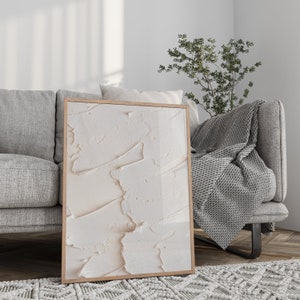 May include: A framed abstract art print with a white and cream textured design. The print is hanging on a white wall above a gray couch with a gray throw blanket.
