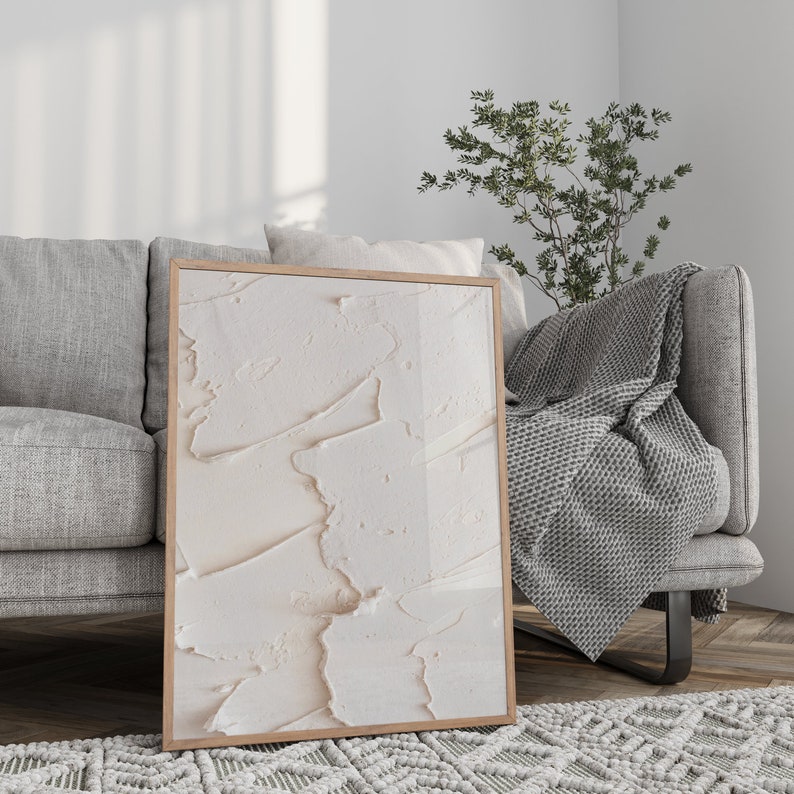 May include: A framed abstract art print with a white and cream textured design. The print is hanging on a white wall above a gray couch with a gray throw blanket.